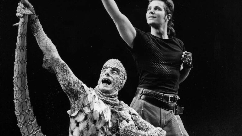 Gabriel Barre in full lizard costume holing his tail up above his head as he sings in a crouched position. Brian Lane Green stands behind him with one arm up in the same direction looking up. The photo is black and white.