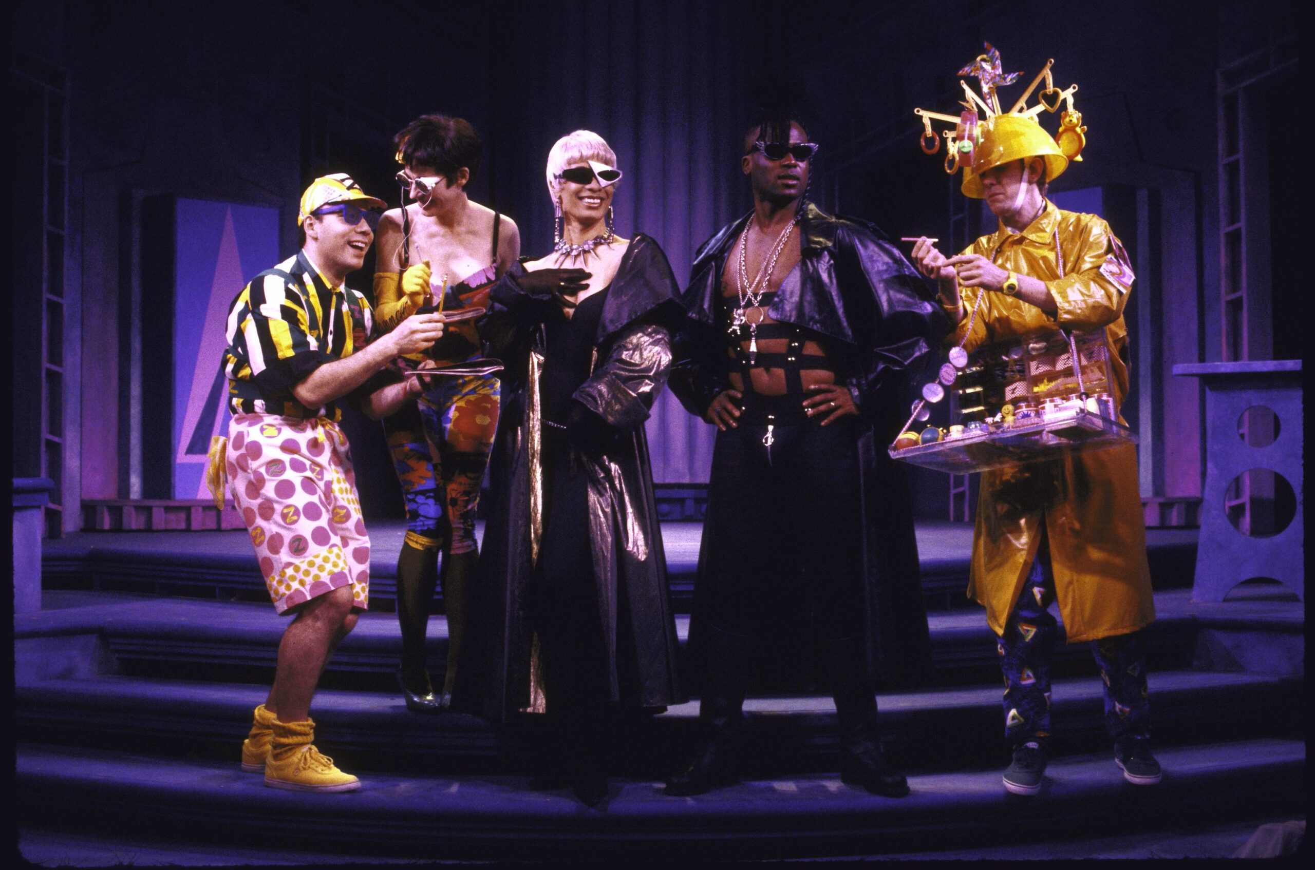 The cast of Weird Romance interact on stage in futuristic costumes holding various props as they perform.