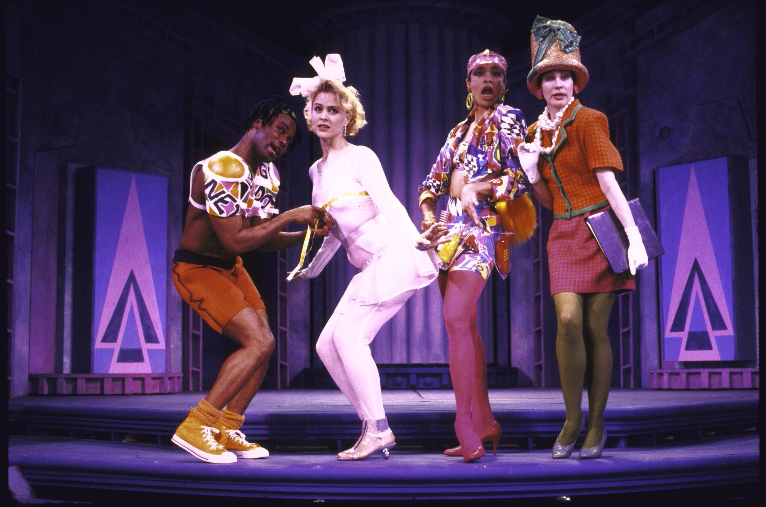 The cast of Weird Romance are dressed in brightly colored costumes with big hats and strong poses as they sing to the audience.