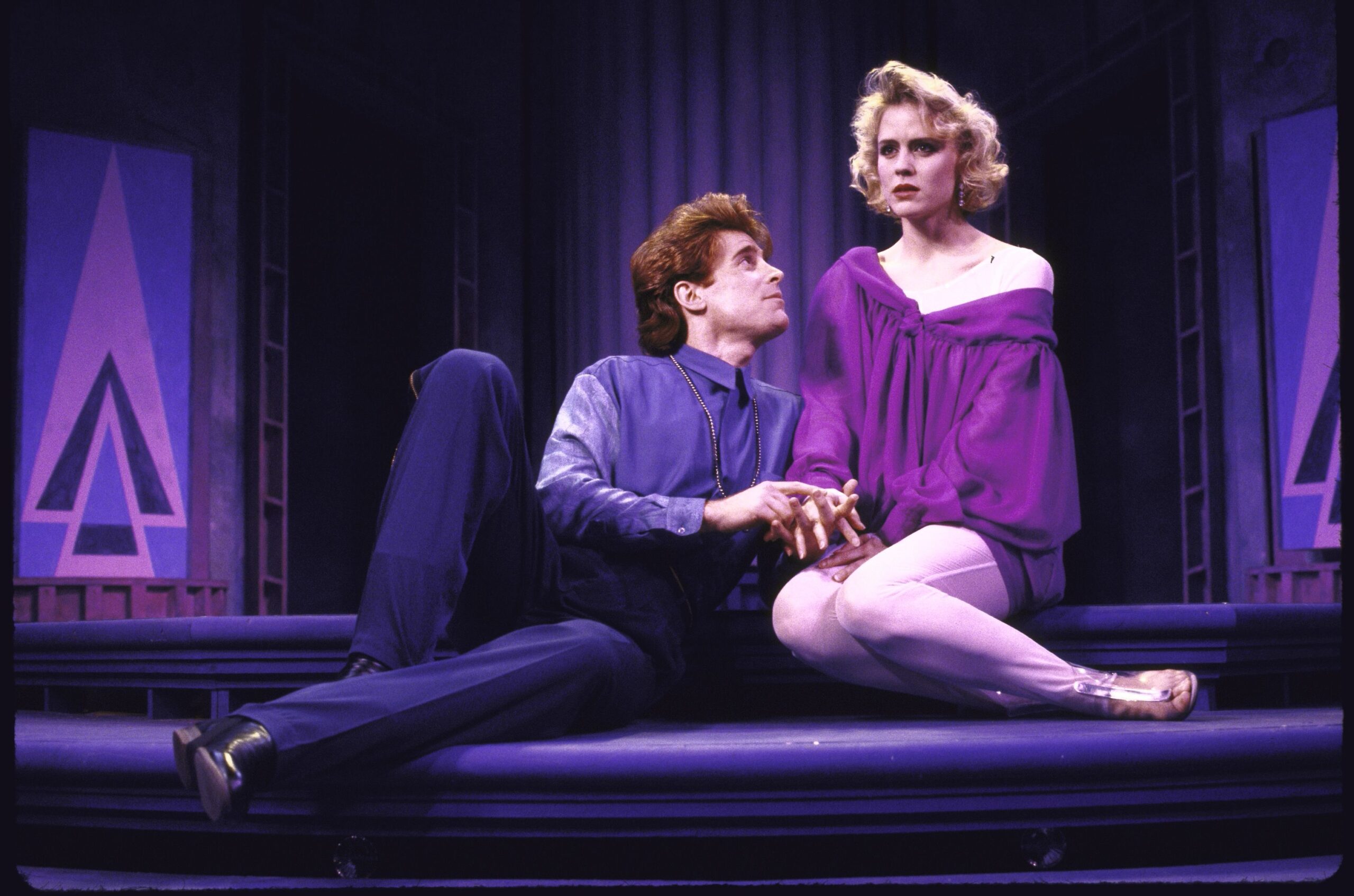 Marguerite McIntyre sits looking out while Sal Viviano sits sort of leaning toward and looking up at her. She is wearing a billowy purple blouse with one exposed shoulder and leggings with silver shoes. He has a blue button up long sleeve shirt and blue dress pants and black dress shoes. Behind them are posters with large purple and black triangular logos. 