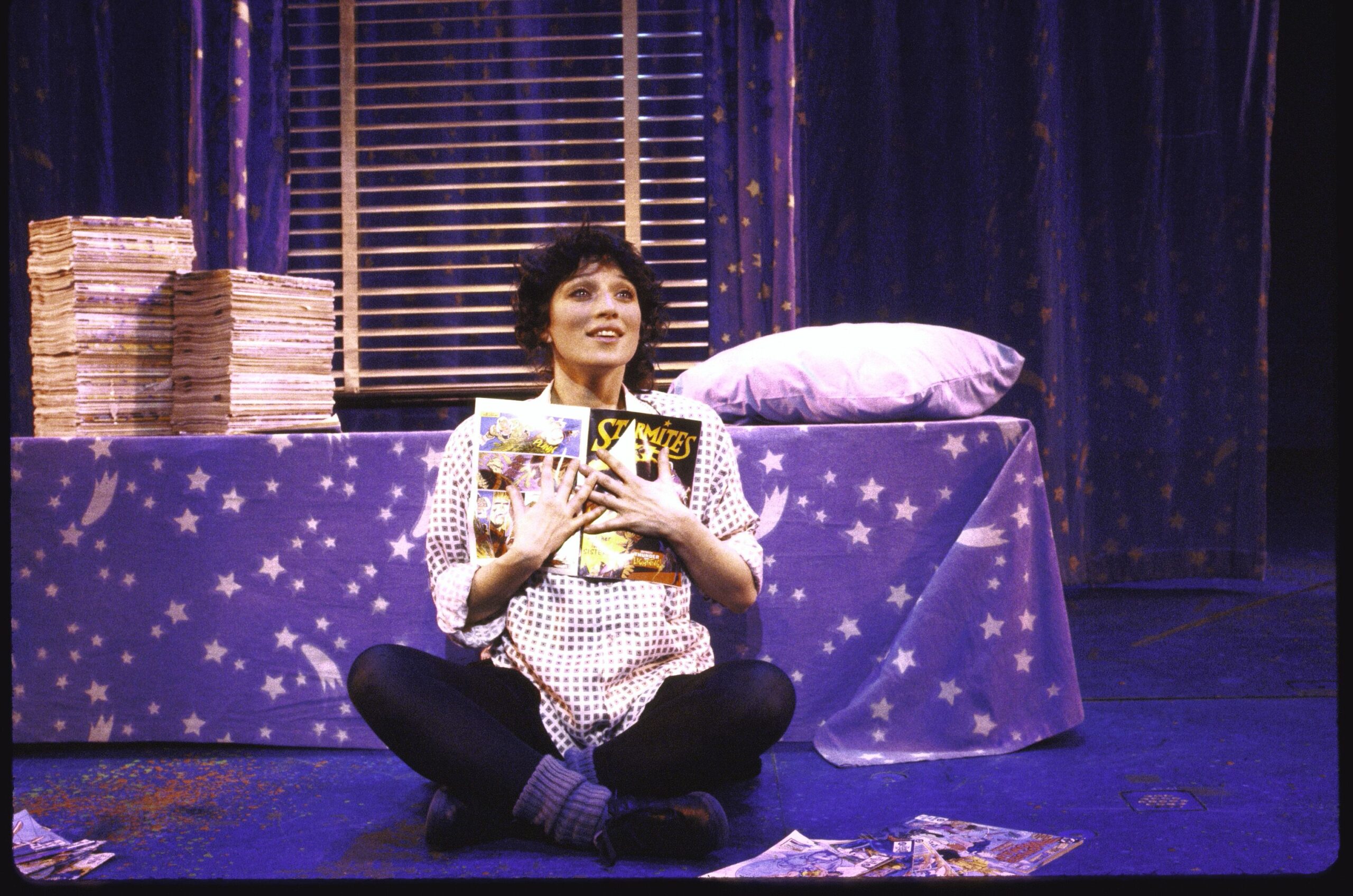 Actress Liz Larsen in a scene from the Broadway musical "Starmites" (New York). A young woman sitting cross-legged holding a comic book called "Starmites" to her chest and looking out longingly with a smile. There is a twin bed behind her with star filled sheets and on top of the bed is a huge pile of comic books. She is on a stage. The bed is part of a set for the musical.