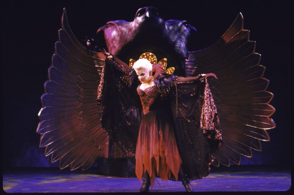 Sharon McNight standing with arms outstretched wearing a fabulous maroon cape and sexy red fairy goddess outfit with high collar and short blonde hair. Behind her is a winged cave/throne.