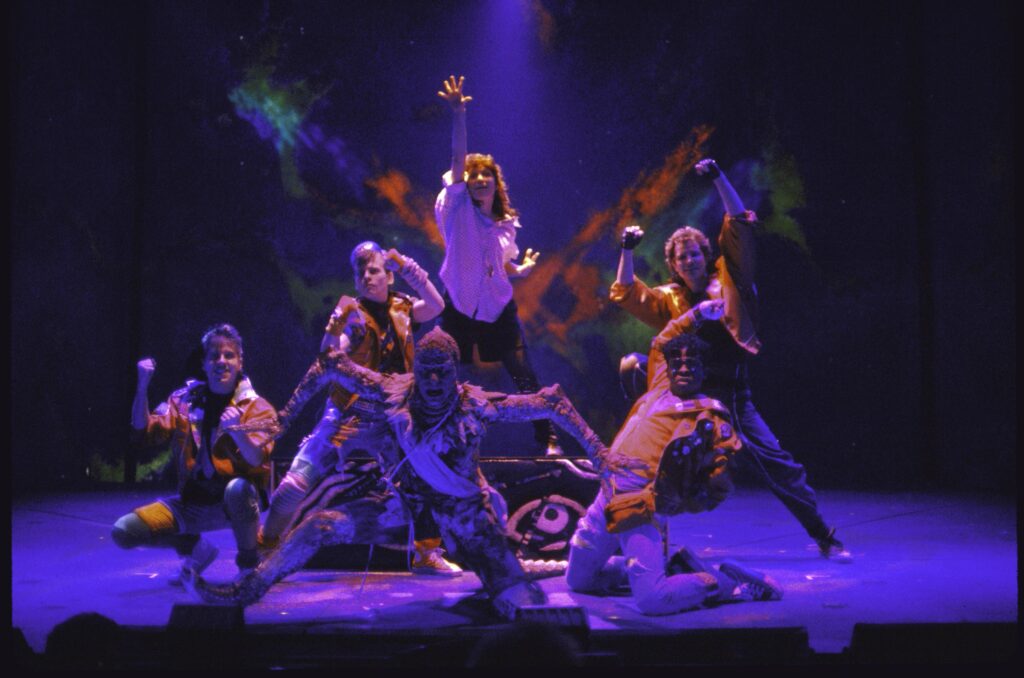 The cast of six characters in various heroic poses like they've just finished a song and the lighting is a dim purplish hue on the stage.