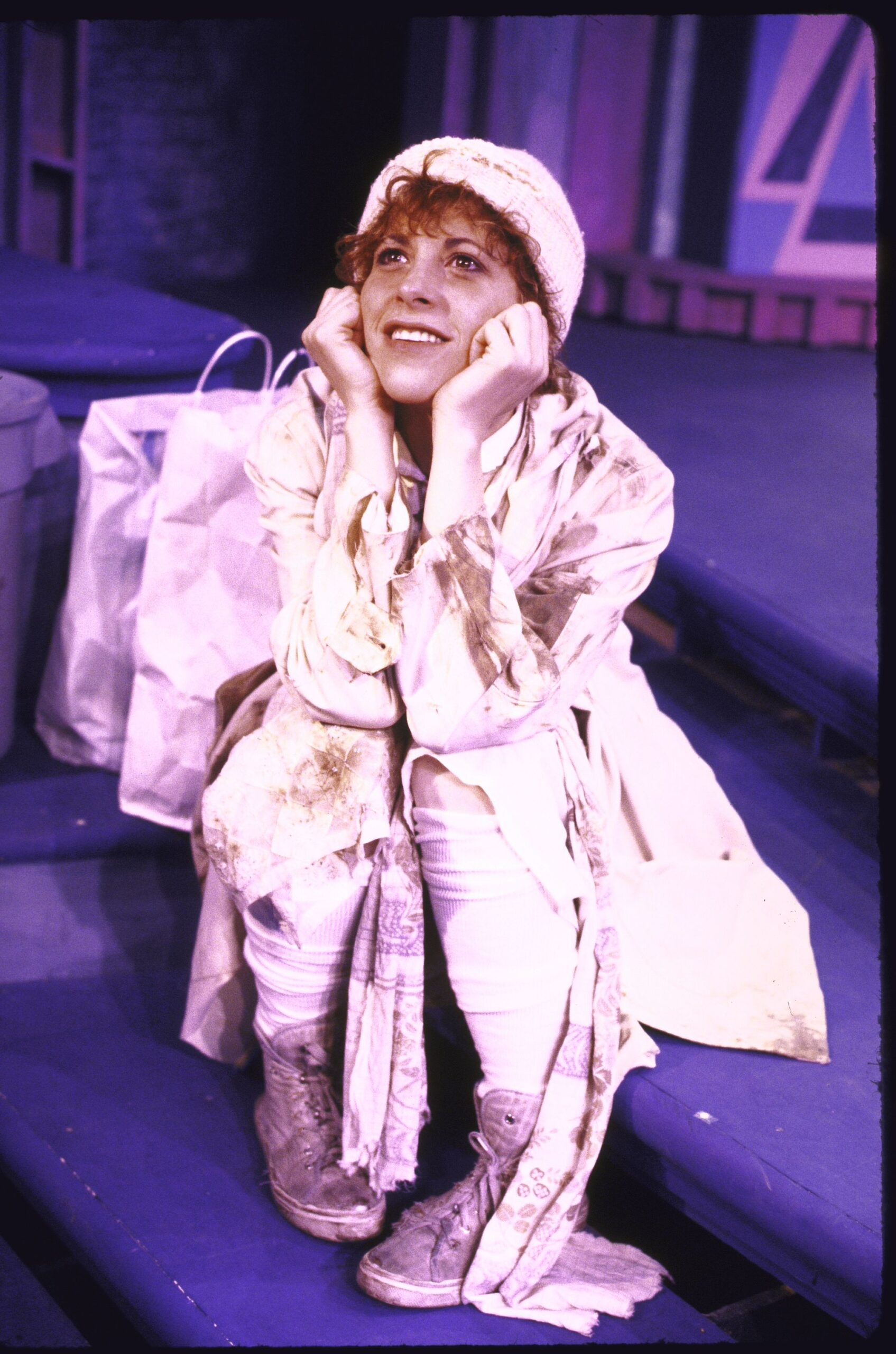 Ellen Greene dressed as a bag lady sits with her head in her hands looking up wistfully.